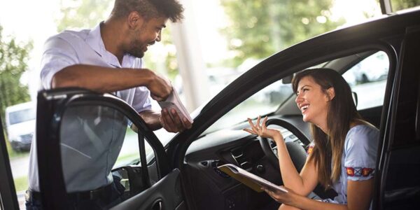 Young woman smiling while taking with a salesperson about payment options for a new car purchase in a car dealership salon. She is sitting in a car.