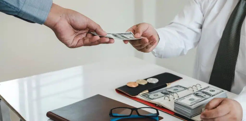 Two hands cup a small stack of US dollar bills close to the chest against a blurred casual background