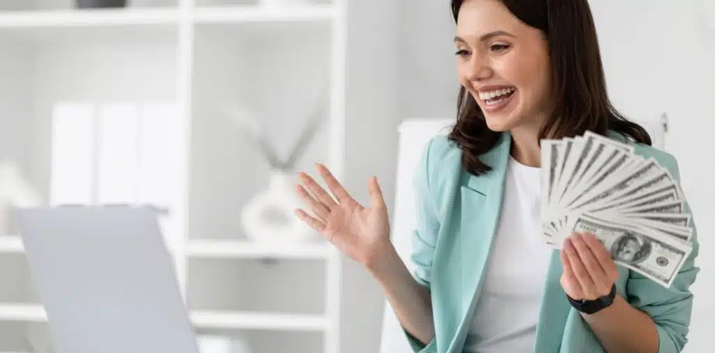 A woman in a pink top inserts a folded dollar bill into a small white ceramic piggy bank on a table
