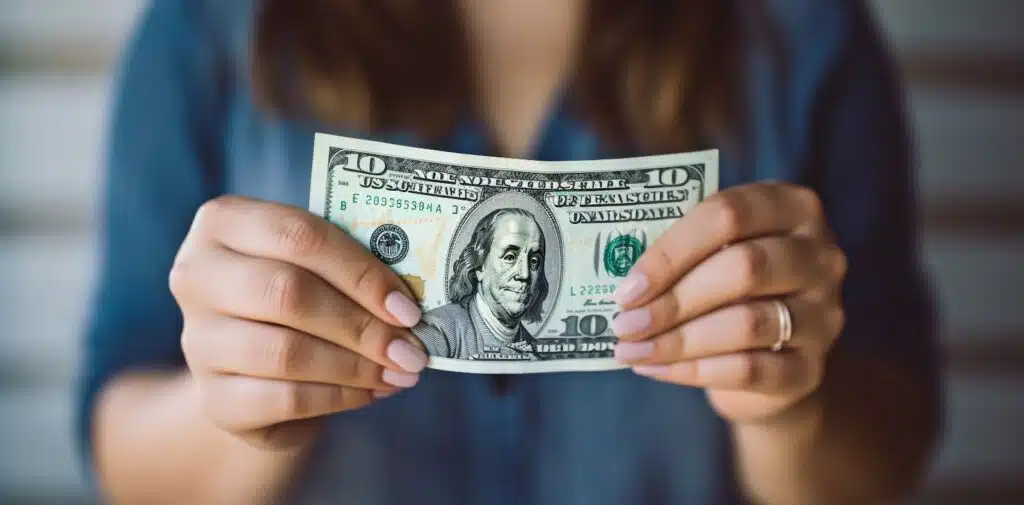 A woman in a patterned jacket holds a folded $100 bill at a desk with a computer monitor and scattered money