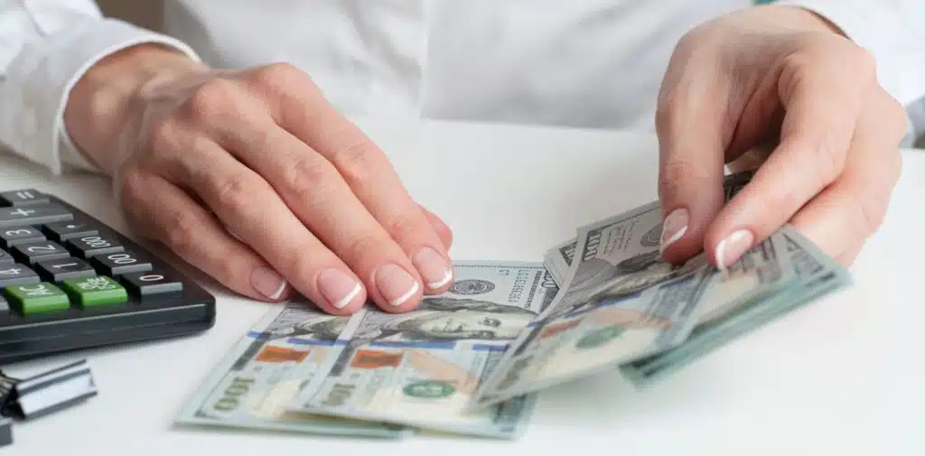 Hands hold a large fan of US $100 bills with a calculator and laptop visible on a wooden desk behind them